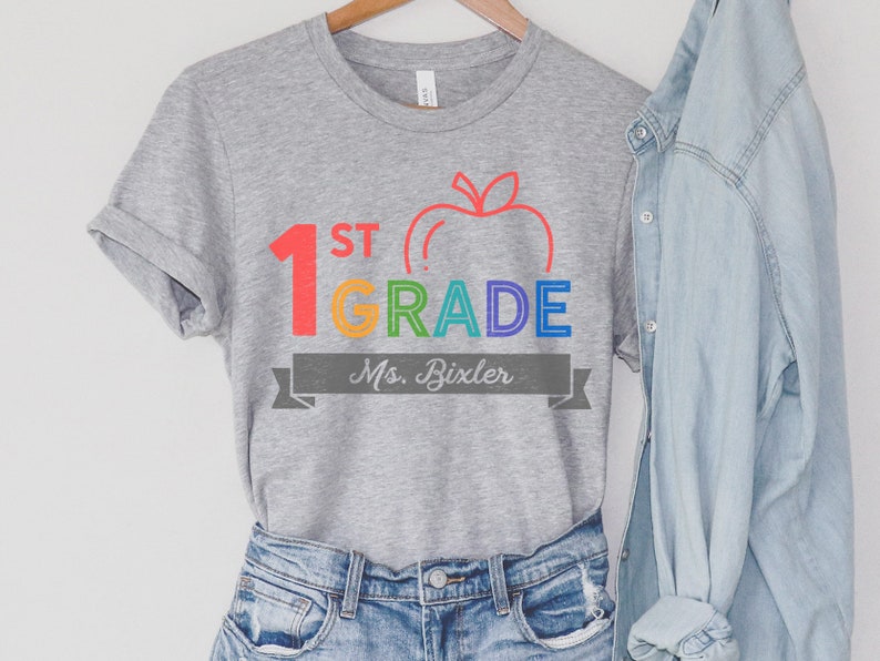 May include: Gray t-shirt with a rainbow colored graphic that says "1st Grade" with an apple and the text "Ms. Bixler" in a gray banner.