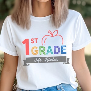 May include: White t-shirt with a rainbow colored "1st Grade" design and a red apple graphic. The text "Ms. Bixler" is printed on a gray banner below the design.