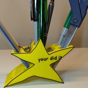 May include: A yellow star-shaped desk organizer with the words "your did it" printed on the front. The organizer has slots for pens, pencils, and scissors.