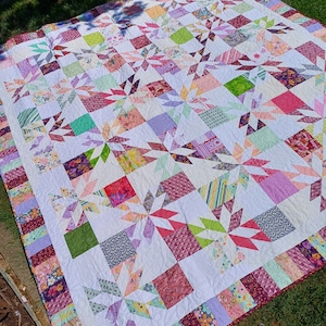 Könnte beinhalten: Eine große Steppdecke mit weißem Hintergrund und einem Patchwork-Muster in verschiedenen Rosatönen, Lila, Grün und Rot. Die Steppdecke besteht aus Quadraten und Dreiecken und die Kanten sind mit einem floralen Stoffrand verziert.