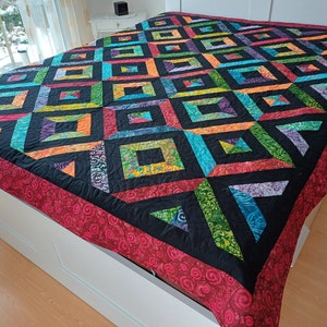 May include: A colorful quilt with a geometric pattern of squares and triangles. The quilt is made with a variety of fabrics in shades of red, orange, yellow, green, blue, purple, and black. The quilt is edged with a red fabric with a swirl pattern.