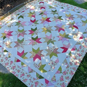 Pode incluir: Um edredom branco com um padrão de estrelas em tons de rosa, verde e amarelo. O edredom tem uma borda floral e está colocado sobre uma superfície gramada.