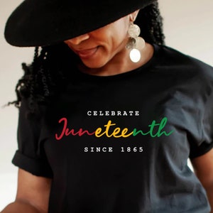 以下が含まれることがあります： 「CELEBRATE Juneteenth SINCE 1865」の文字が赤、黄、緑で書かれた黒いTシャツ。Tシャツを着ている人は黒い帽子と大きなイヤリングをしています。
