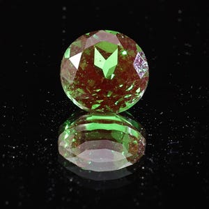 Pode incluir: Uma gema redonda e facetada com uma mistura de tons verdes e castanho-avermelhados. A gema está sobre um fundo preto, com o seu reflexo por baixo. As facetas da gema capturam a luz, criando um efeito cintilante.