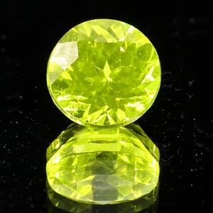 May include: A round, faceted, bright lime-green gemstone is centered on a reflective black surface. The gemstone's facets catch the light, creating a sparkling effect. The gem's reflection is visible below it.