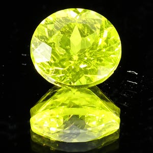 May include: A faceted, round, bright yellow-green gemstone sits on a reflective black surface. The gemstone's facets catch the light, creating a sparkling effect. The gem is a vibrant, eye-catching piece.