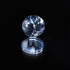 May include: A round, faceted, light blue gemstone with a brilliant cut, set against a black background. The gem's reflection is visible below it, creating a symmetrical image. The gemstone is likely a precious stone.