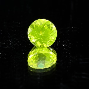 May include: A round, faceted, bright lime green gemstone is displayed against a black background. The gem is reflected in the glossy surface below, creating a symmetrical image. The gemstone's facets catch the light, showcasing its brilliance.