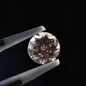 Könnte beinhalten: Ein runder, funkelnder Diamant, gehalten von einer Pinzette vor schwarzem Hintergrund. Die Facetten des Diamanten reflektieren das Licht und erzeugen ein brillantes Farbspiel. Die Pinzette ist hellgrau.