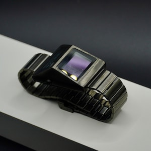 May include: A black digital watch with a metal band. The watch face is rectangular and has a dark purple display.