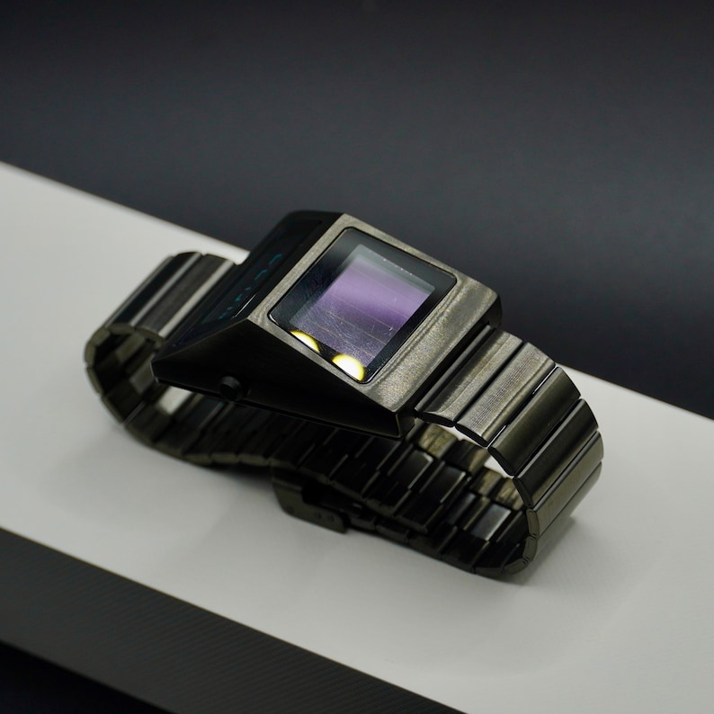 May include: A black digital watch with a metal band. The watch face is rectangular and has a dark purple display.