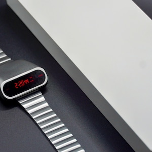 Op de afbeelding: Een zilveren digitale horloge met een zwarte wijzerplaat en een rood digitaal display dat de tijd 14:20:44 laat zien. Het horloge ligt op een zwart oppervlak.
