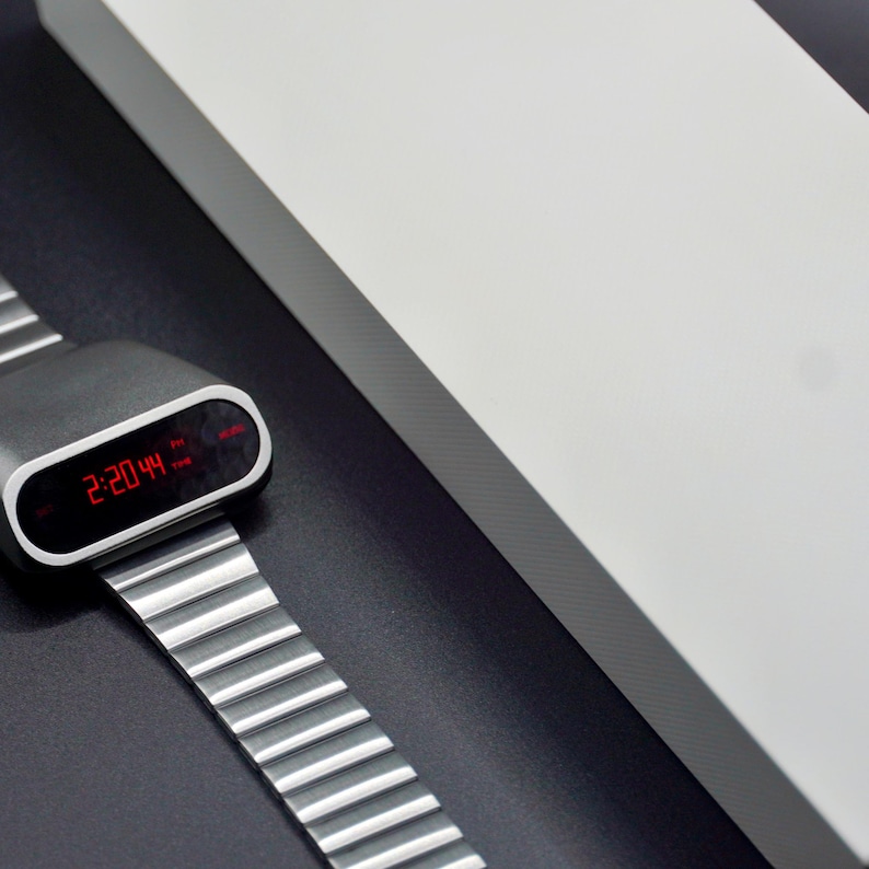 Op de afbeelding: Een zilveren digitale horloge met een zwarte wijzerplaat en een rood digitaal display dat de tijd 14:20:44 laat zien. Het horloge ligt op een zwart oppervlak.