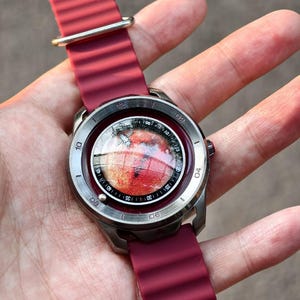 May include: A wristwatch with a burgundy silicone band and a silver-colored case. The watch face displays a red and orange image, possibly a planet or celestial body. The watch has a round face with white numerals.