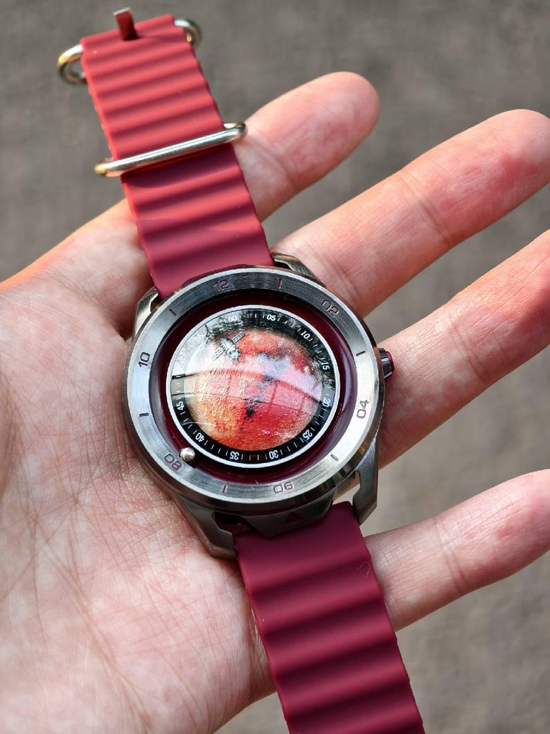 May include: A wristwatch with a burgundy silicone band and a silver-colored case. The watch face displays a red and orange image, possibly a planet or celestial body. The watch has a round face with white numerals.