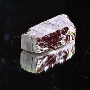 Könnte beinhalten: Ein rechteckiger, roher Edelstein mit tiefroter Farbe, der Licht reflektiert. Der Stein hat eine raue, strukturierte Oberfläche mit sichtbaren kristallinen Strukturen. Das Bild ist vor einem schwarzen Hintergrund platziert, wobei sich der Edelstein unten spiegelt.