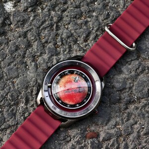 May include: A wristwatch with a burgundy silicone strap and a silver-toned case. The watch face features a red and white design, possibly depicting a planet. The watch is set against a textured gray surface.