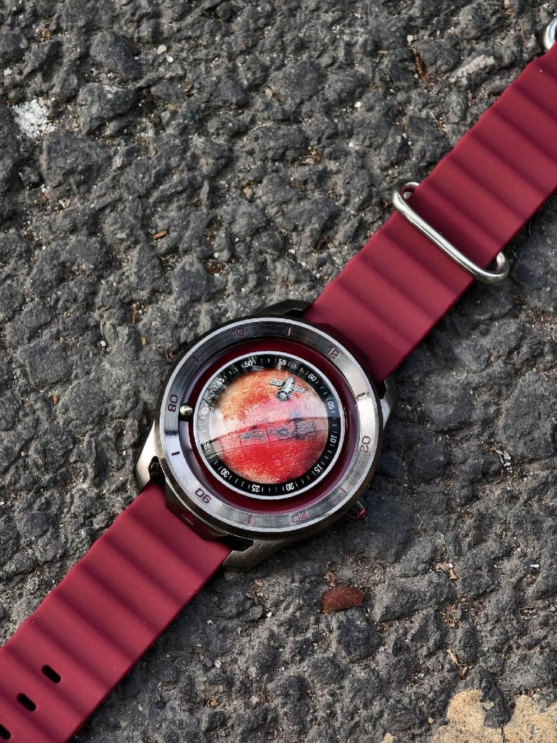 May include: A wristwatch with a burgundy silicone strap and a silver-toned case. The watch face features a red and white design, possibly depicting a planet. The watch is set against a textured gray surface.