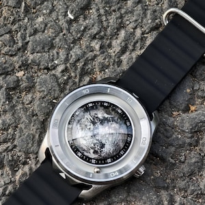 May include: A silver-toned wristwatch with a black band. The watch face features a detailed, grayscale image, possibly a celestial body. The watch is set against a textured, dark gray surface.