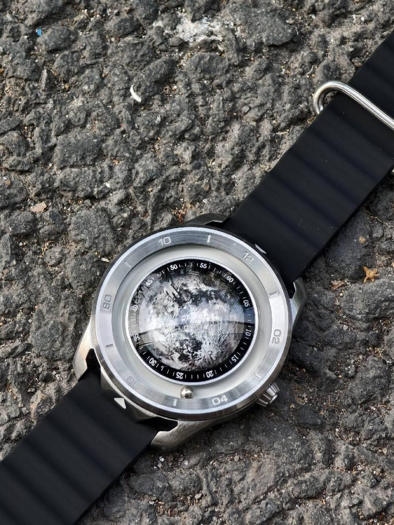 May include: A silver-toned wristwatch with a black band. The watch face features a detailed, grayscale image, possibly a celestial body. The watch is set against a textured, dark gray surface.