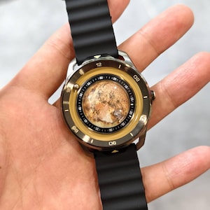 May include: A wristwatch with a black ribbed band and a silver-colored case. The watch face has a unique design with a brown and tan planet-like image in the center, surrounded by a gold-colored ring with black numerals. The watch is held by a hand.