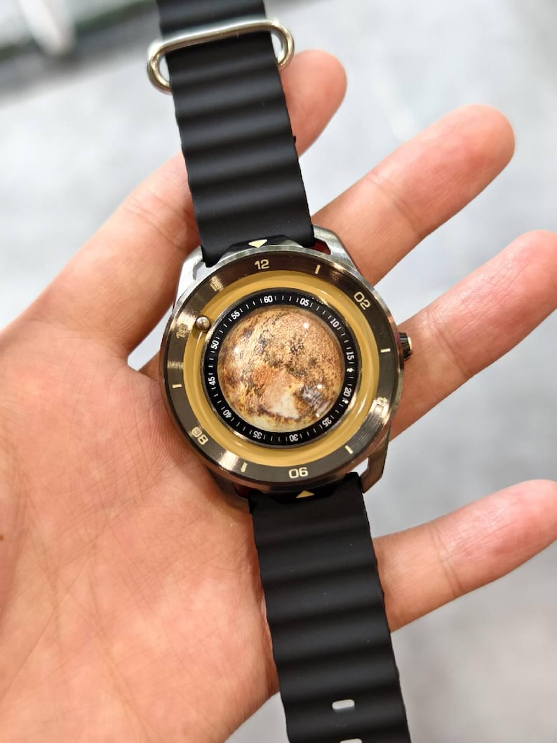 May include: A wristwatch with a black ribbed band and a silver-colored case. The watch face has a unique design with a brown and tan planet-like image in the center, surrounded by a gold-colored ring with black numerals. The watch is held by a hand.