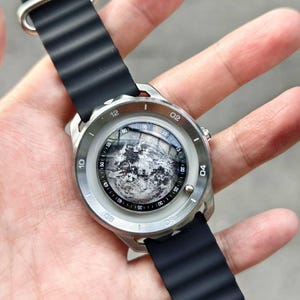 May include: A silver-toned wristwatch with a black rubber band. The watch face features a detailed grayscale image of the moon. The watch has a round face with white numerals and markers. The band is ribbed.