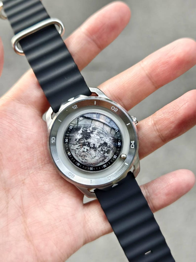 May include: A silver-toned wristwatch with a black rubber band. The watch face features a detailed grayscale image of the moon. The watch has a round face with white numerals and markers. The band is ribbed.