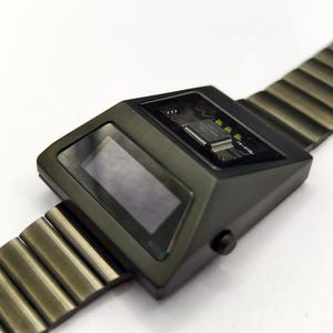 Puede incluir: Un reloj de pulsera digital negro con una banda de metal. La esfera del reloj es rectangular y tiene una pantalla digital. El reloj tiene un circuito impreso visible.