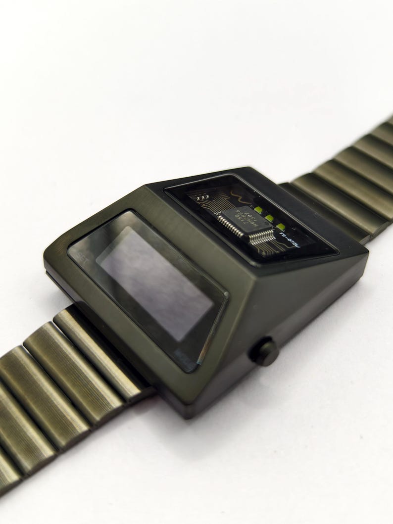 Puede incluir: Un reloj de pulsera digital negro con una banda de metal. La esfera del reloj es rectangular y tiene una pantalla digital. El reloj tiene un circuito impreso visible.