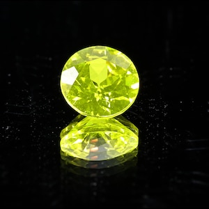 May include: A round, faceted, bright yellow-green gemstone, possibly peridot, is centered against a black background. The gem's facets reflect light, creating a sparkling effect. The gem's reflection is visible below.