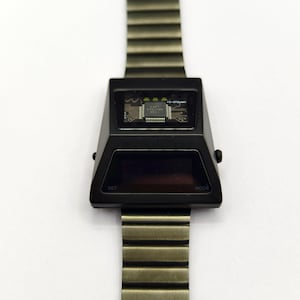 Puede incluir: Un reloj de pulsera digital negro con una correa de metal plateado. La esfera del reloj es una pantalla rectangular negra con las palabras "SET" y "MODE" impresas en las esquinas inferiores izquierda y derecha. La esfera del reloj tambi&eacute;n tiene una peque&ntilde;a ventana que muestra una placa de circuito con el texto "FL-073MC" impreso en ella.