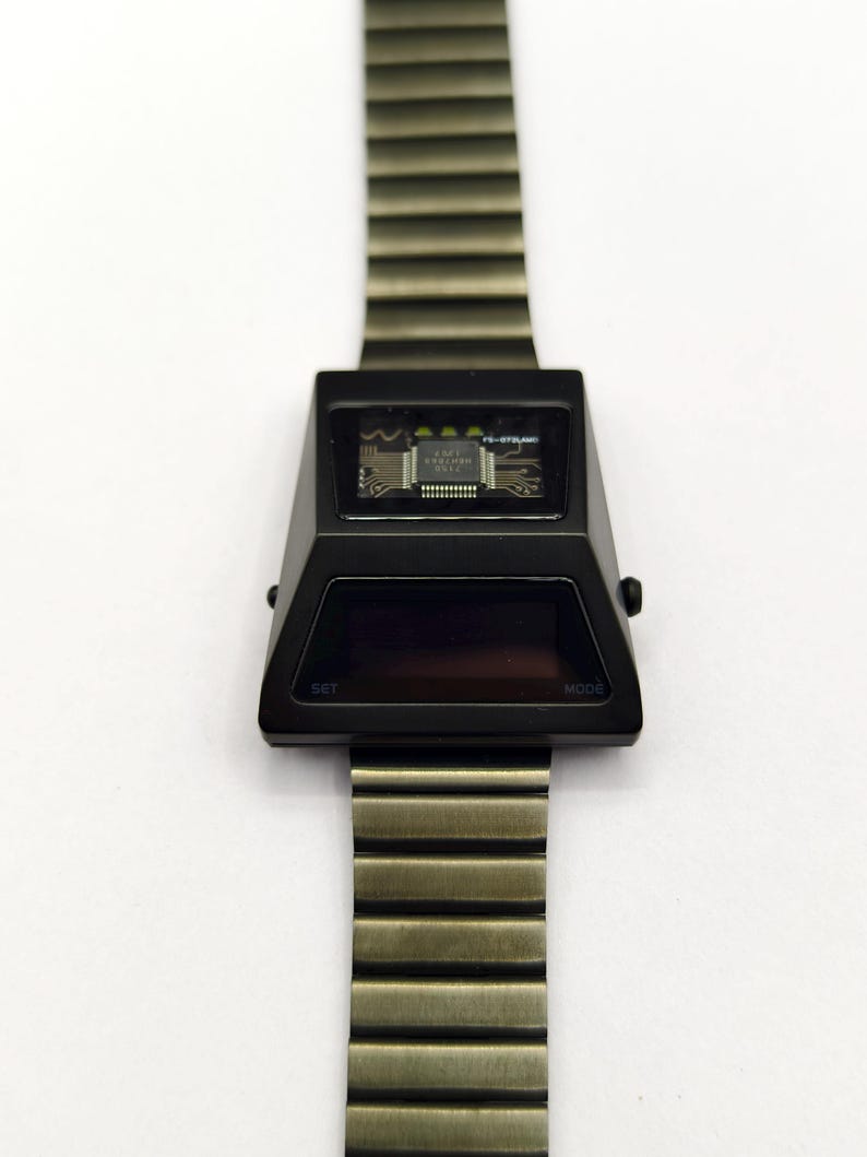 Puede incluir: Un reloj de pulsera digital negro con una correa de metal plateado. La esfera del reloj es una pantalla rectangular negra con las palabras "SET" y "MODE" impresas en las esquinas inferiores izquierda y derecha. La esfera del reloj tambi&eacute;n tiene una peque&ntilde;a ventana que muestra una placa de circuito con el texto "FL-073MC" impreso en ella.