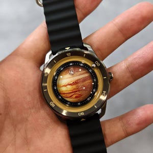 May include: A wristwatch with a black band and a round face featuring a detailed image of Jupiter. The watch has a silver-colored case and a black rubber strap. The watch face displays the time with white numerals.