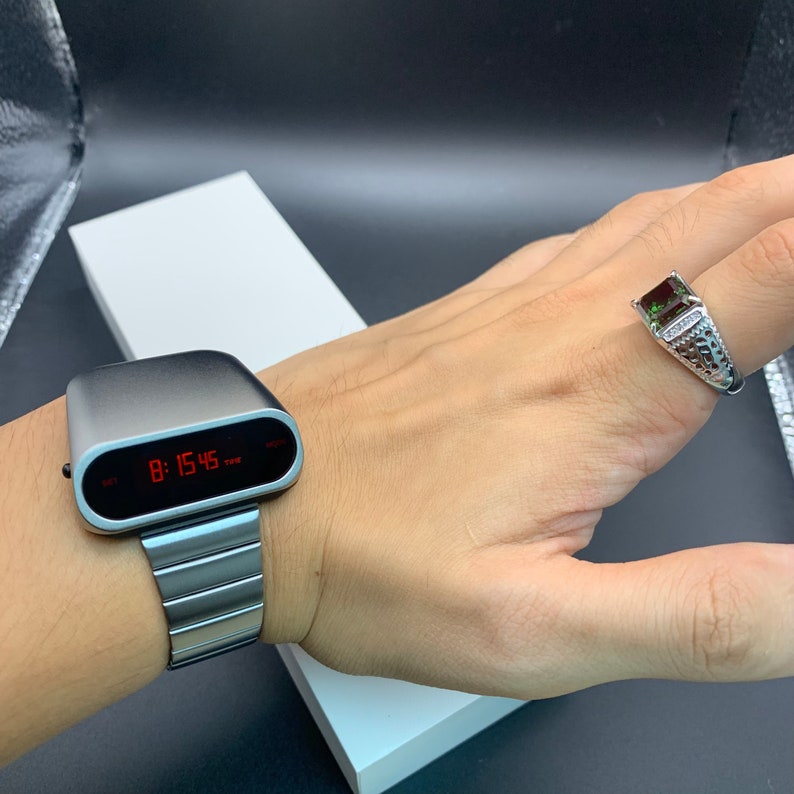 Op de afbeelding: Een zilveren digitaal horloge met een rood digitaal display dat de tijd 8:15:45 laat zien. Het horloge zit om de pols van een persoon. De persoon draagt ook een zilveren ring met een groene edelsteen.