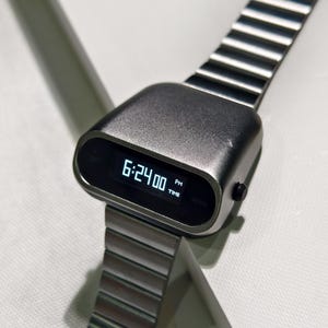 Op de afbeelding: Een zilveren digitale polshorloge met een zwart display dat de tijd 18:24:00 uur weergeeft. Het horloge heeft een uniek, afgerond, futuristisch ontwerp en een zilveren metalen band.