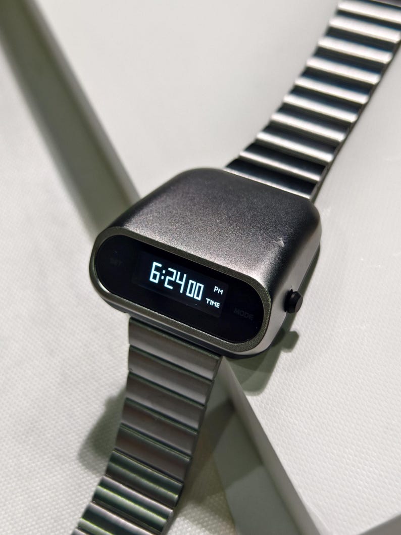 Op de afbeelding: Een zilveren digitale polshorloge met een zwart display dat de tijd 18:24:00 uur weergeeft. Het horloge heeft een uniek, afgerond, futuristisch ontwerp en een zilveren metalen band.