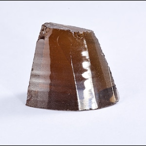 Könnte beinhalten: Ein brauner, dreieckiger Kristall mit rauer, strukturierter Oberfläche. Der Kristall hat eine polierte, reflektierende Seite, die das Licht einfängt und einen Kontrast zwischen glatten und rauen Texturen erzeugt. Der Kristall liegt auf weißem Hintergrund.