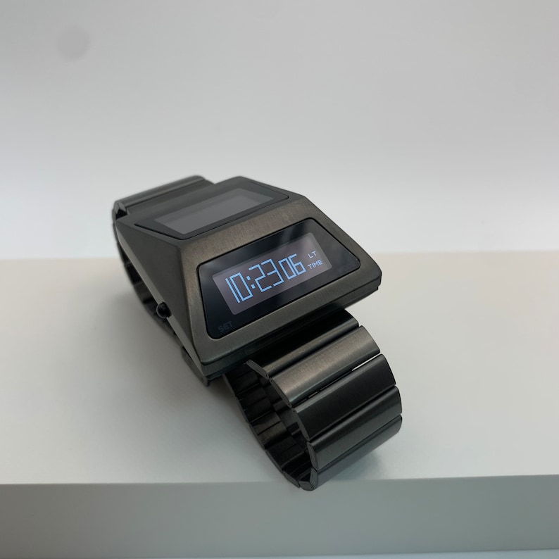 May include: A black digital wristwatch with a rectangular face and a metal band. The watch face displays the time as 10:23:06.