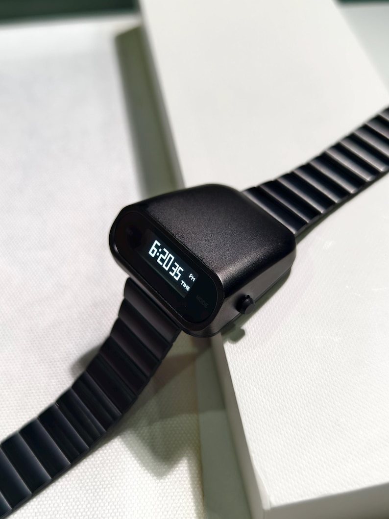 Op de afbeelding: Een zwart digitaal polshorloge met een zwarte metalen band. Het horloge laat de tijd 18:20:35 zien.