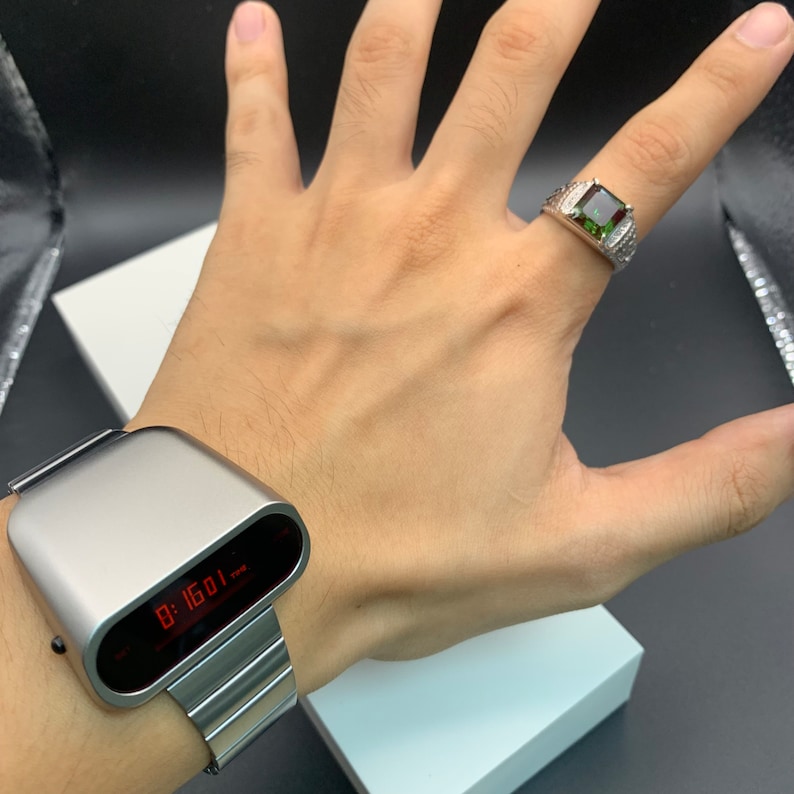 Op de afbeelding: Een zilveren digitaal horloge met een rood digitaal display dat de tijd 8:16:01 laat zien. Het horloge zit om de pols van een persoon, en aan de andere hand zit een zilveren ring met een groene edelsteen.