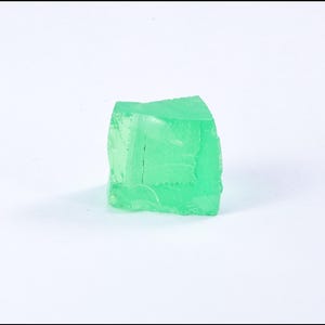 Könnte beinhalten: Ein einzelnes, durchscheinendes, hellgrünes, würfelförmiges Objekt mit einer rauen, unebenen Oberfläche. Das Objekt befindet sich vor einem schlichten weißen Hintergrund, der seine Farbe und Form hervorhebt. Der Würfel scheint fest zu sein, möglicherweise ein dekorativer Gegenstand.