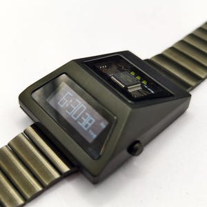 Puede incluir: Un reloj de pulsera digital negro con una banda de metal. La esfera del reloj muestra la hora como "8E:0E" y tiene una peque&ntilde;a etiqueta "TIME". El reloj tiene un circuito impreso visible.