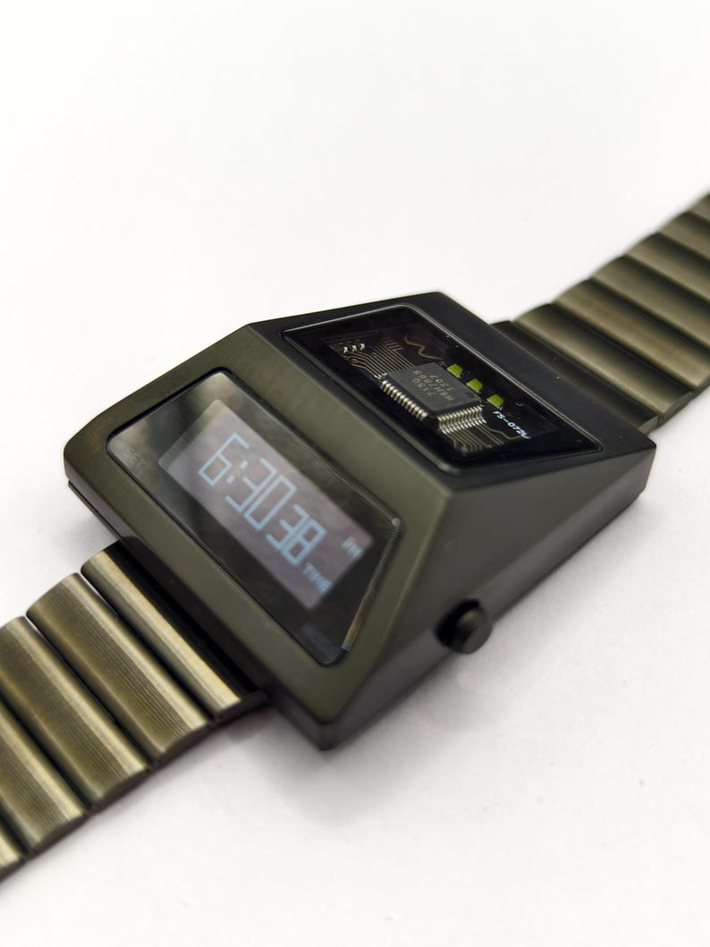 Puede incluir: Un reloj de pulsera digital negro con una banda de metal. La esfera del reloj muestra la hora como "8E:0E" y tiene una peque&ntilde;a etiqueta "TIME". El reloj tiene un circuito impreso visible.