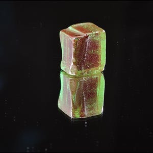 May include: A translucent, cube-shaped gemstone with a mix of red and green hues. The gem is reflected on a glossy black surface, creating a mirrored effect. The cube's edges are slightly irregular, giving it a natural appearance.