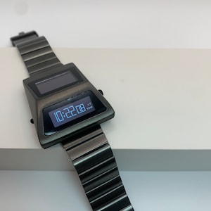 May include: A black digital wristwatch with a silver display showing the time 10:22:08. The watch has a black metal band and a rectangular face.