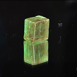 May include: A translucent, square-shaped object with a green hue and brown inclusions. The object is resting on a reflective black surface, creating a mirrored effect. The overall appearance suggests a gemstone or decorative element.