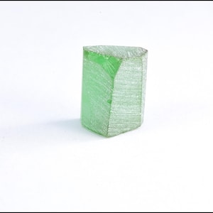 Könnte beinhalten: Ein durchscheinender, hellgrüner Kristall mit einer facettierten, rechteckigen Form. Der Kristall hat eine leicht raue Textur und eine dreieckige Oberseite. Das Objekt ist vor einem schlichten weißen Hintergrund platziert.