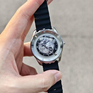 May include: A silver-toned wristwatch with a black rubber band. The watch face features a detailed, grayscale image. The band is ribbed and secured with a silver clasp. The watch is held up against a blurred background.