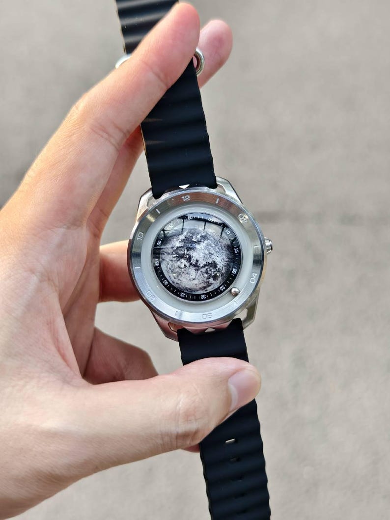May include: A silver-toned wristwatch with a black rubber band. The watch face features a detailed, grayscale image. The band is ribbed and secured with a silver clasp. The watch is held up against a blurred background.